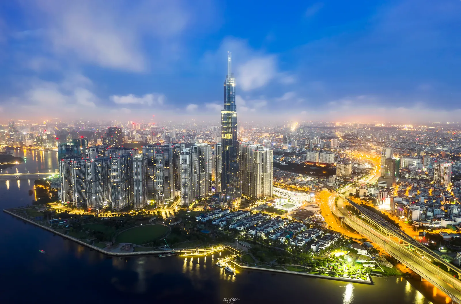 Night view of a very tall tower above a lit riverside park and a curving expressway