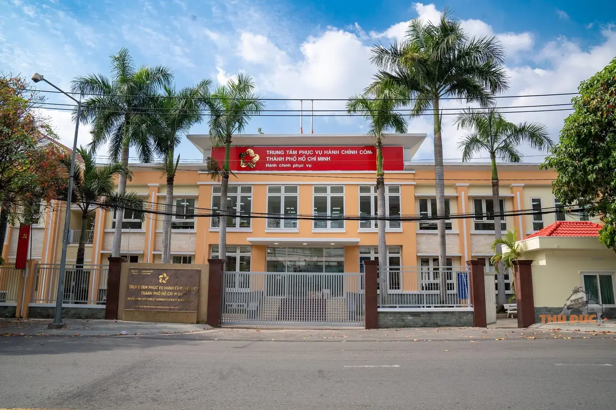 Front of a pale orange public building behind a gate, with palm trees and a red sign above the entrance.