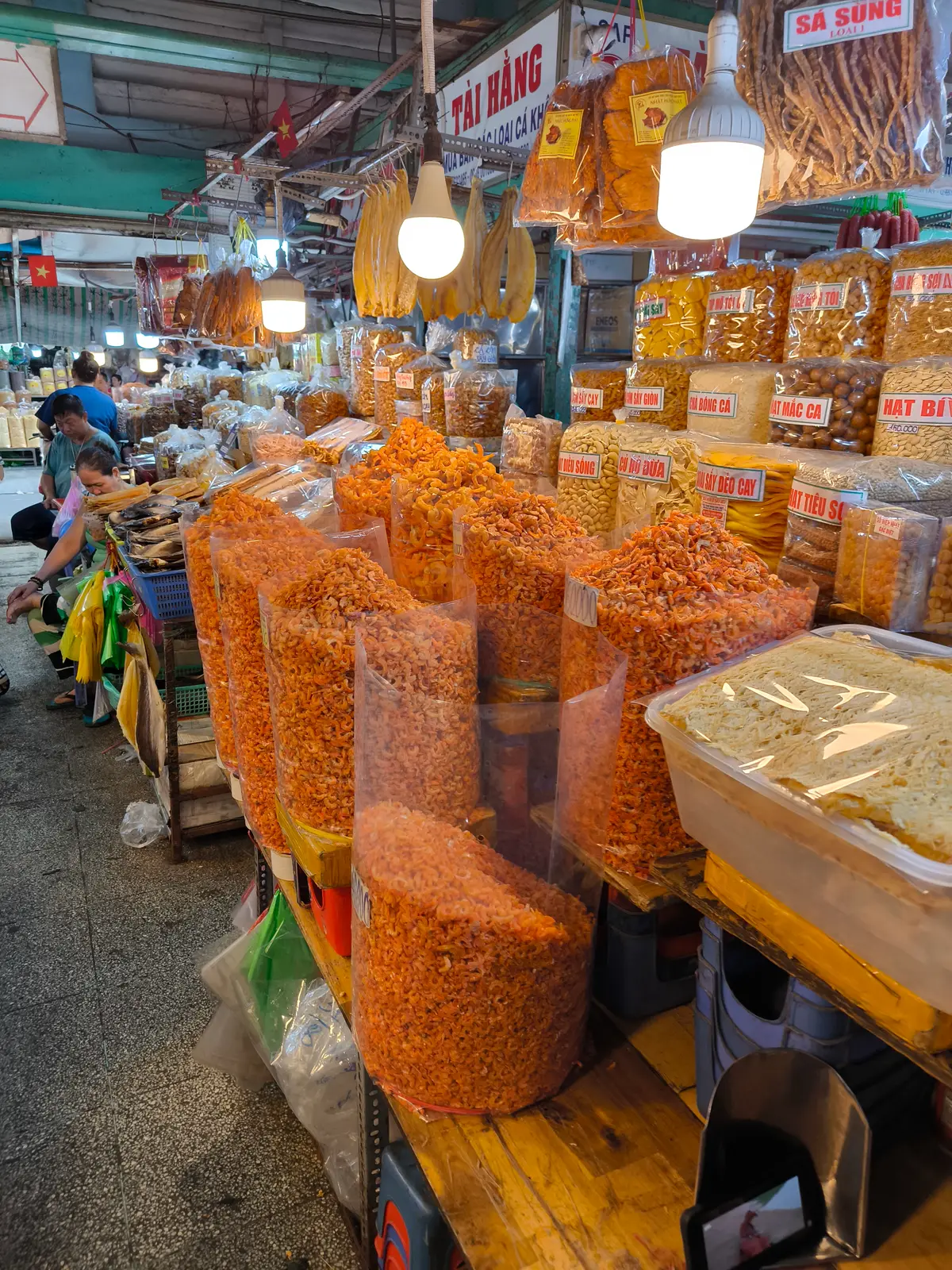 Bags of dried orange shrimp stacked on a wooden stall in a market aisle with hanging lamps