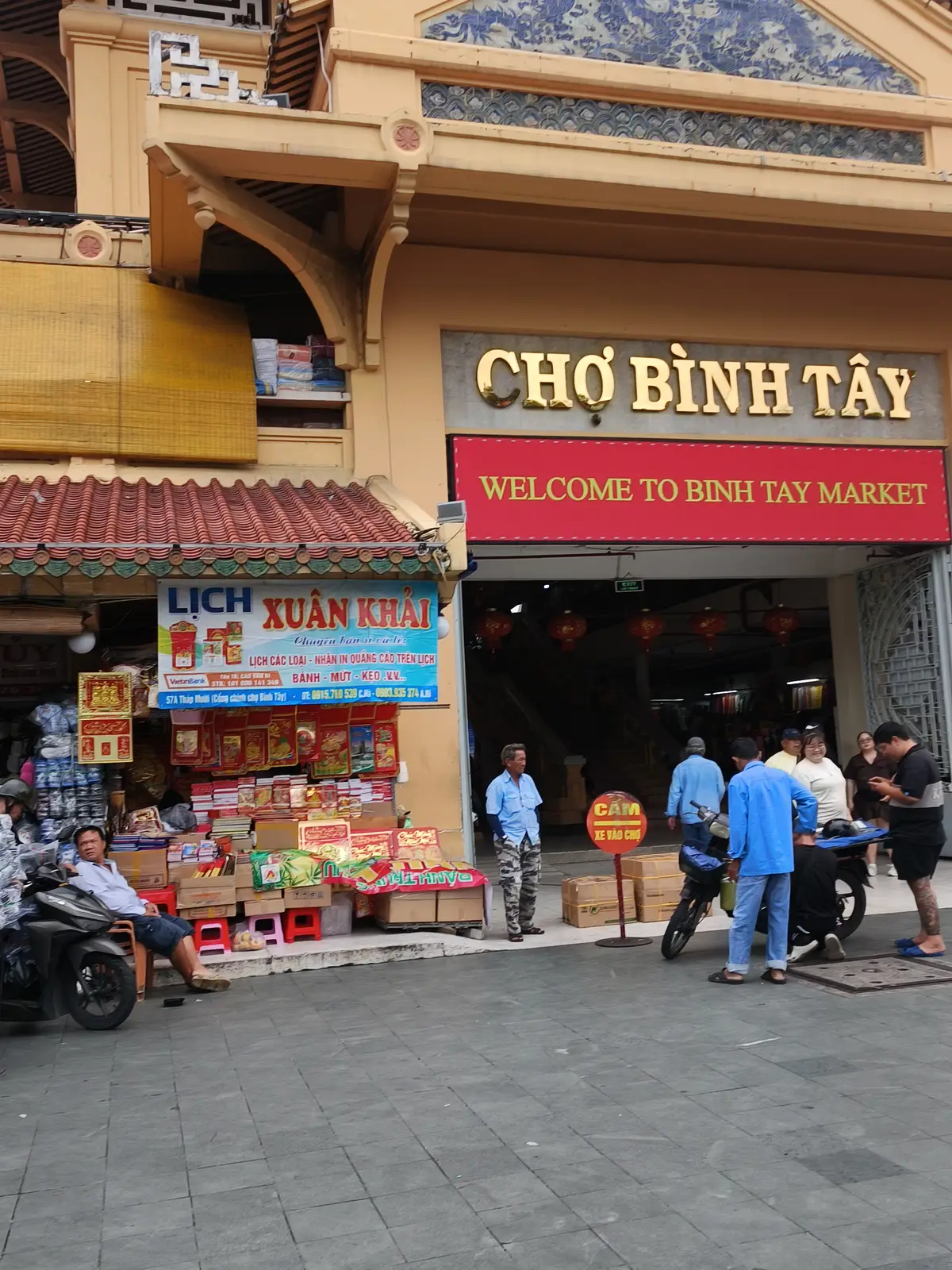 Street entrance under a Cho Binh Tay sign with a welcome banner, a motorbike and shoppers