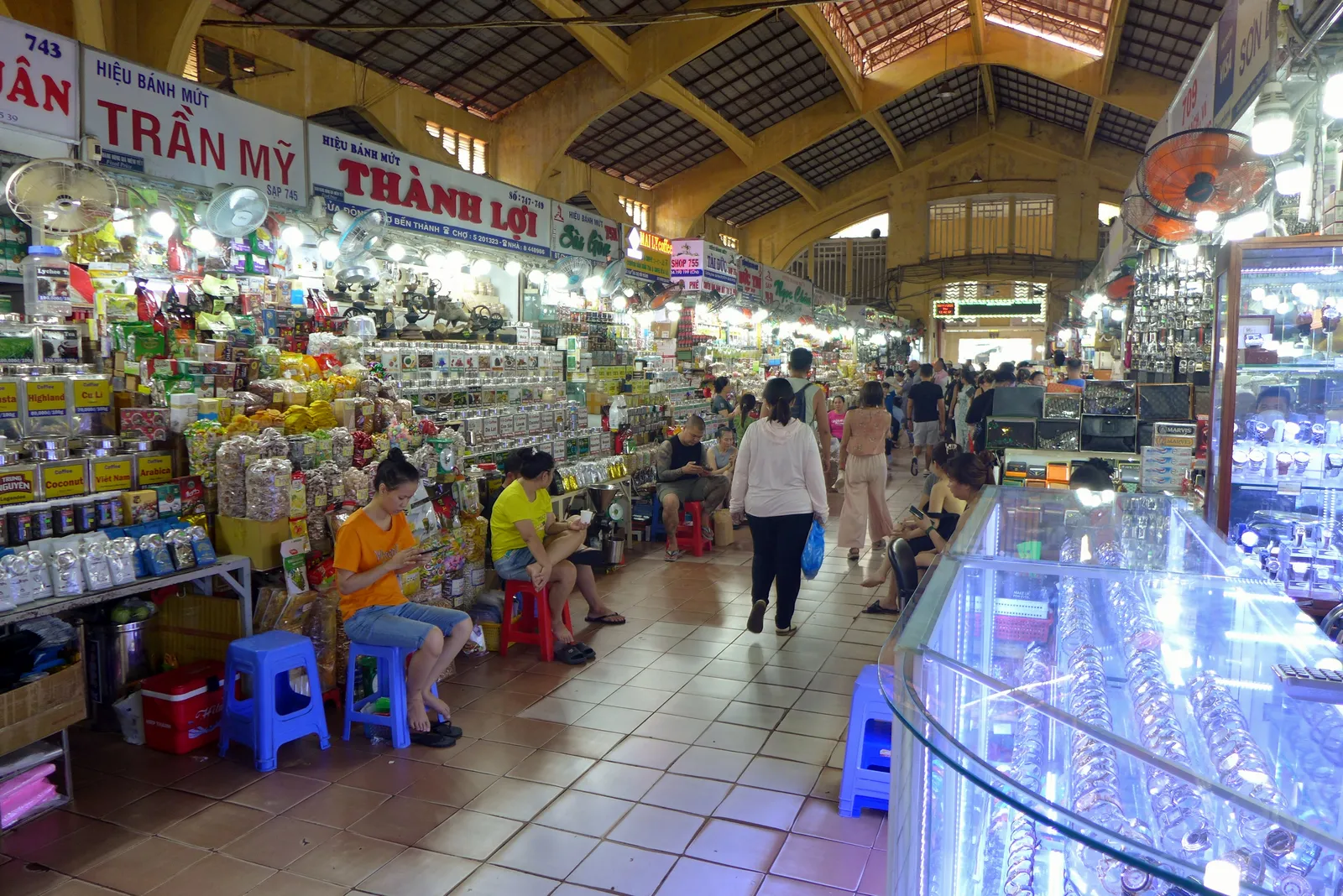 Covered market hall with stalls, shoppers and plastic stools under a high roof