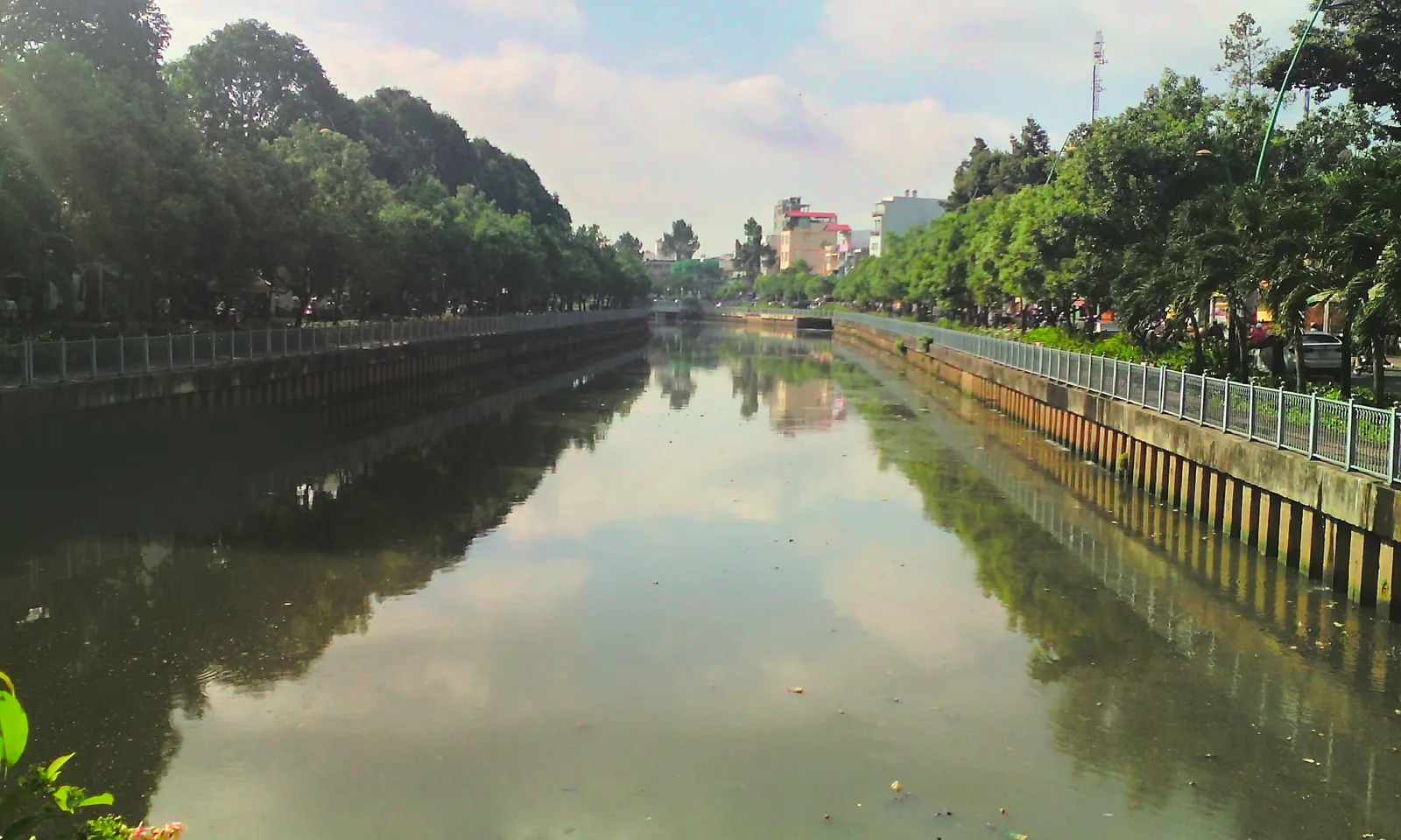 Straight canal lined with trees and a railing on both banks, reflecting the sky