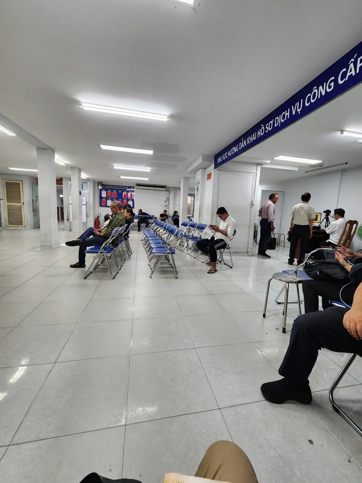Large tiled waiting hall with rows of blue chairs, a few people seated and counters along the far wall.