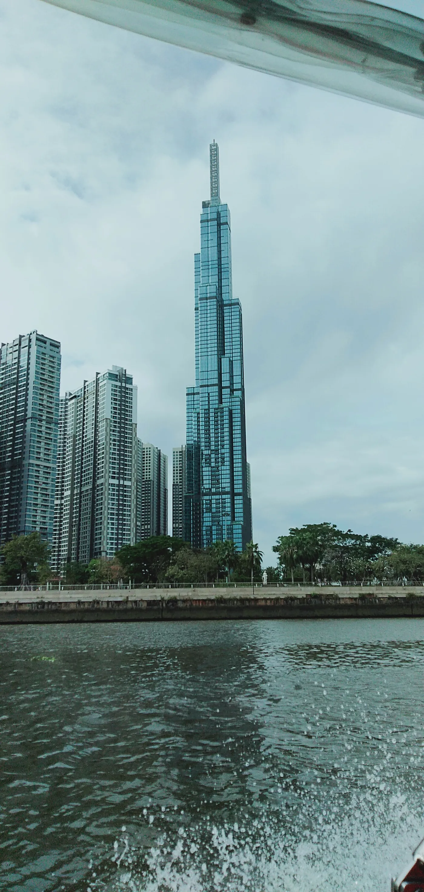 Tall glass tower rising behind riverside apartment blocks, seen across the water from a boat