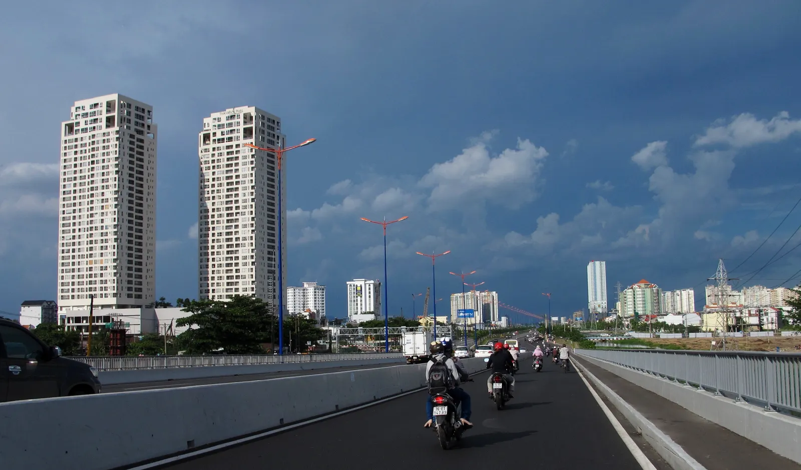 Two tall apartment towers beside a wide road bridge with motorbikes in the foreground under a cloudy sky