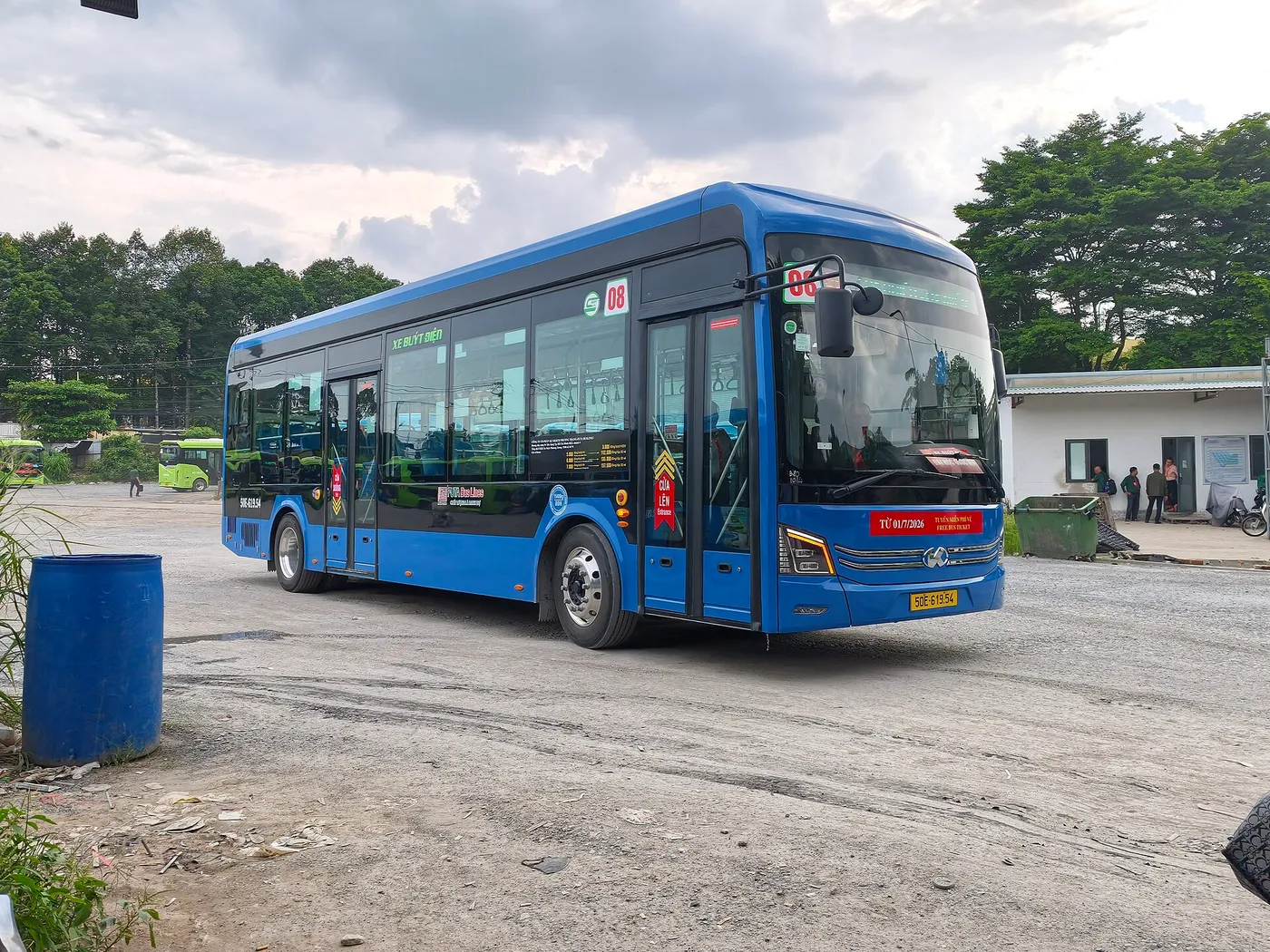 Blue electric city bus parked on an open lot with trees behind