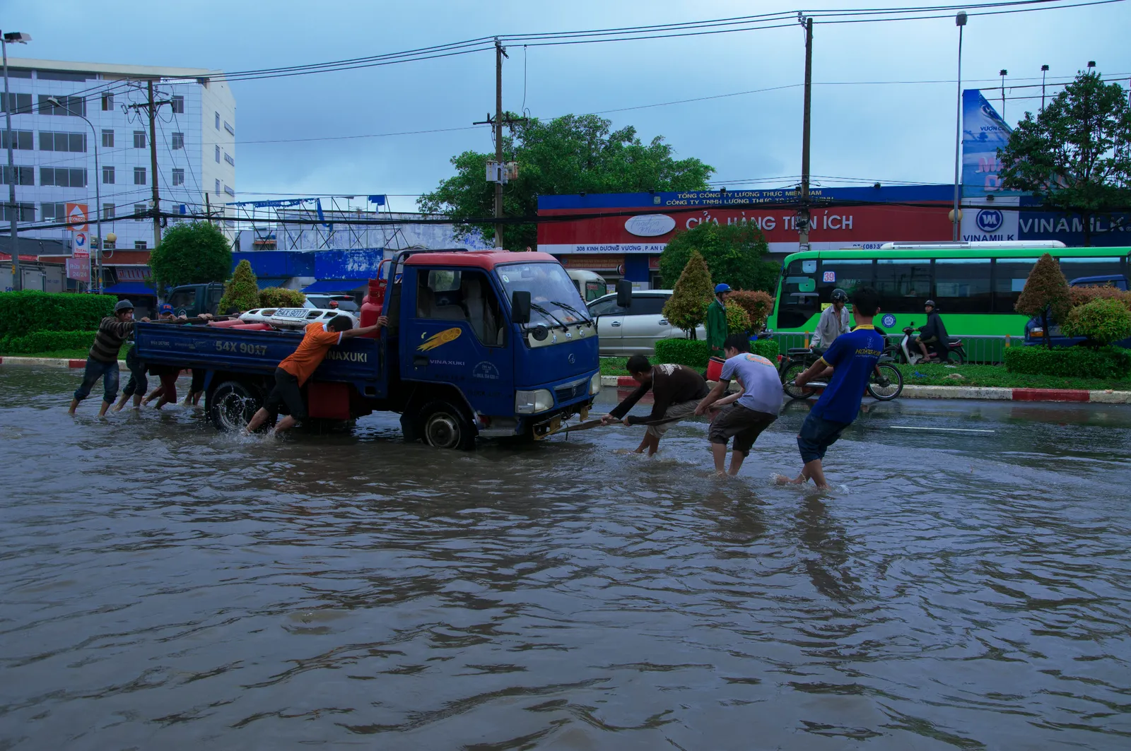 Men pushing a blue truck through a flooded road with a green bus behind
