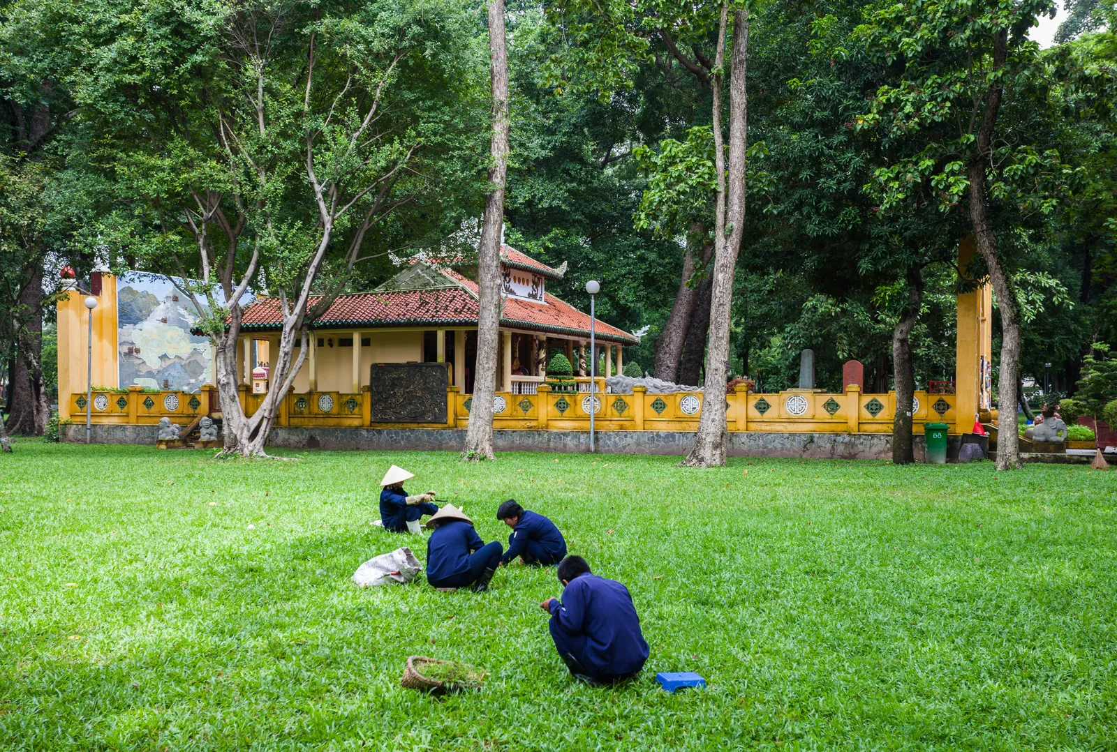 Three people in dark clothes sitting on a green lawn in front of a small yellow temple pavilion among tall trees