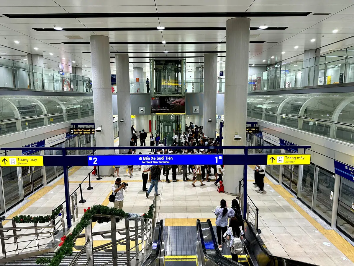 Station concourse with a blue direction sign above two platforms, escalators and a festive garland on the stair rail