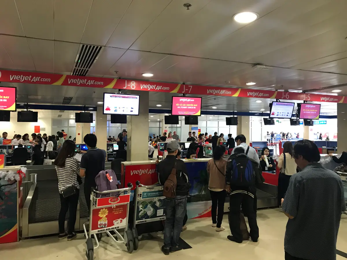 Check-in counters under red airline signs with travellers queuing and luggage trolleys in the foreground