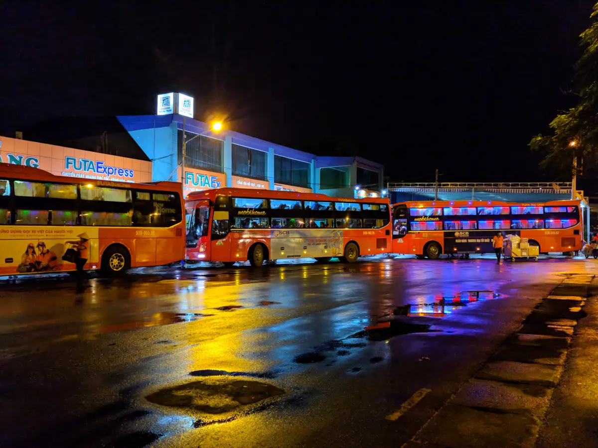 Orange coaches parked in a lit bay at night beside a building with a blue sign