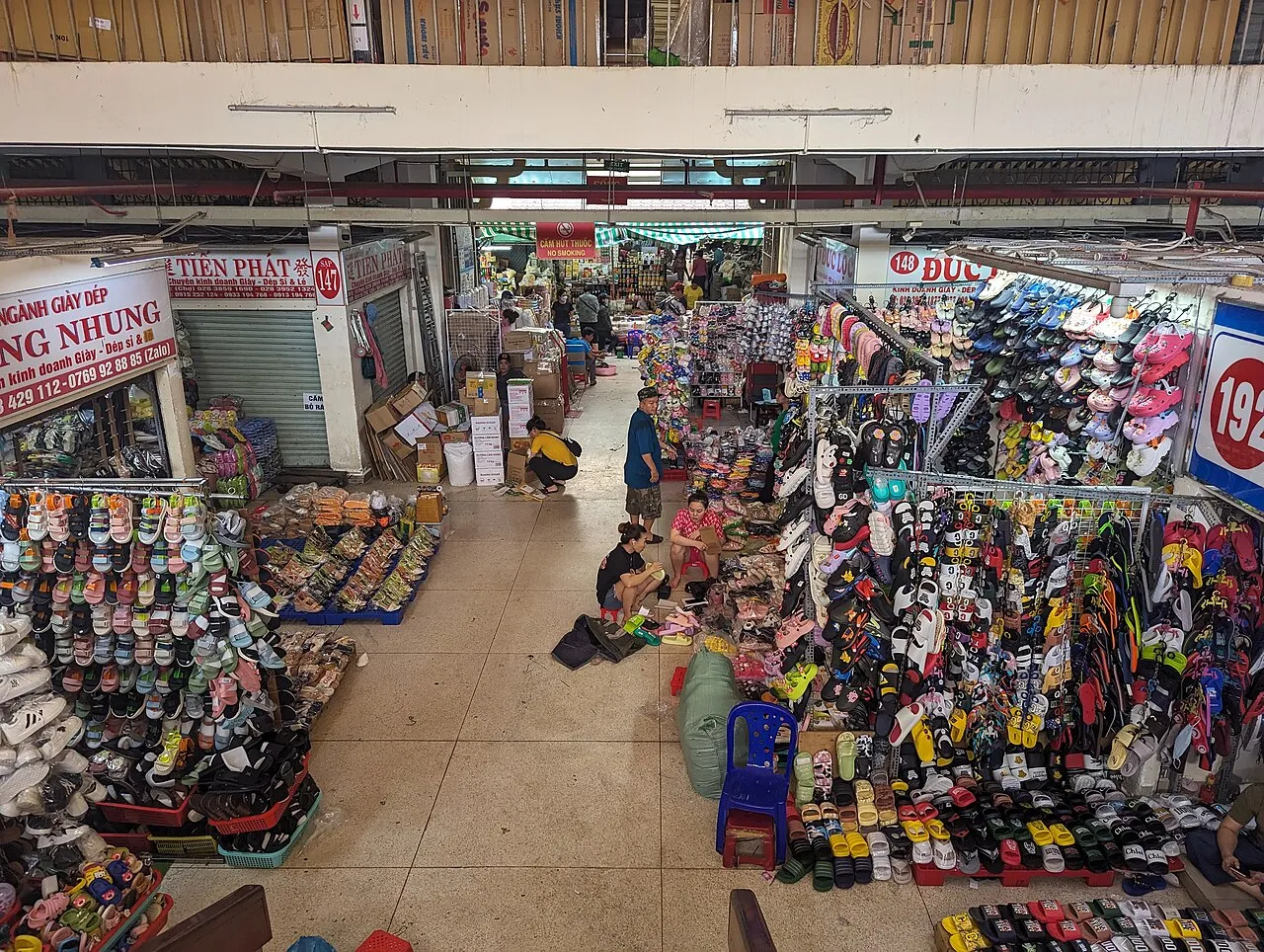 Wide market aisle between stalls selling rows of sandals and shoes, seen from a balcony