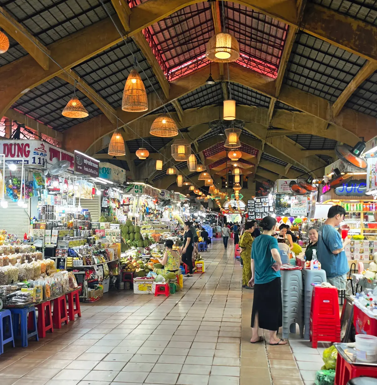 Covered market hall with hanging woven lamps and fruit stalls along a wide aisle