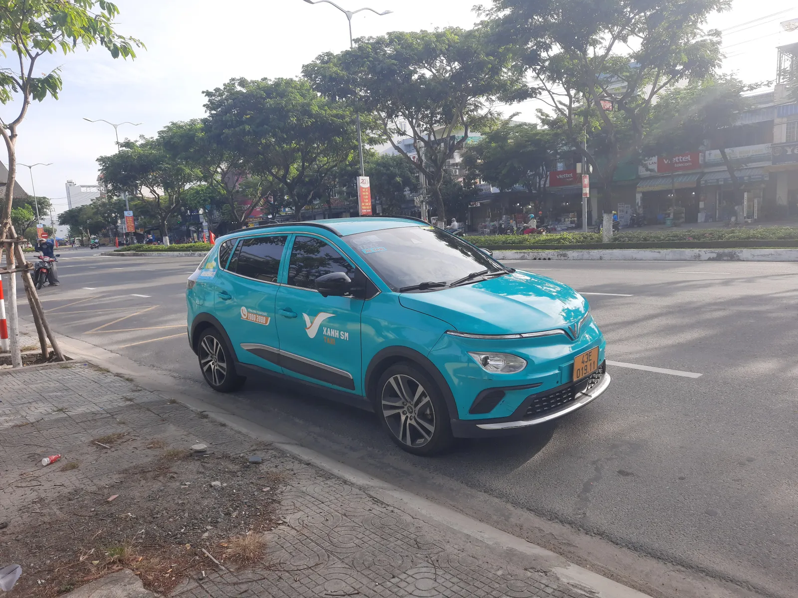 Teal-green compact electric car with a Xanh SM sign on its roof parked beside a tree-lined road