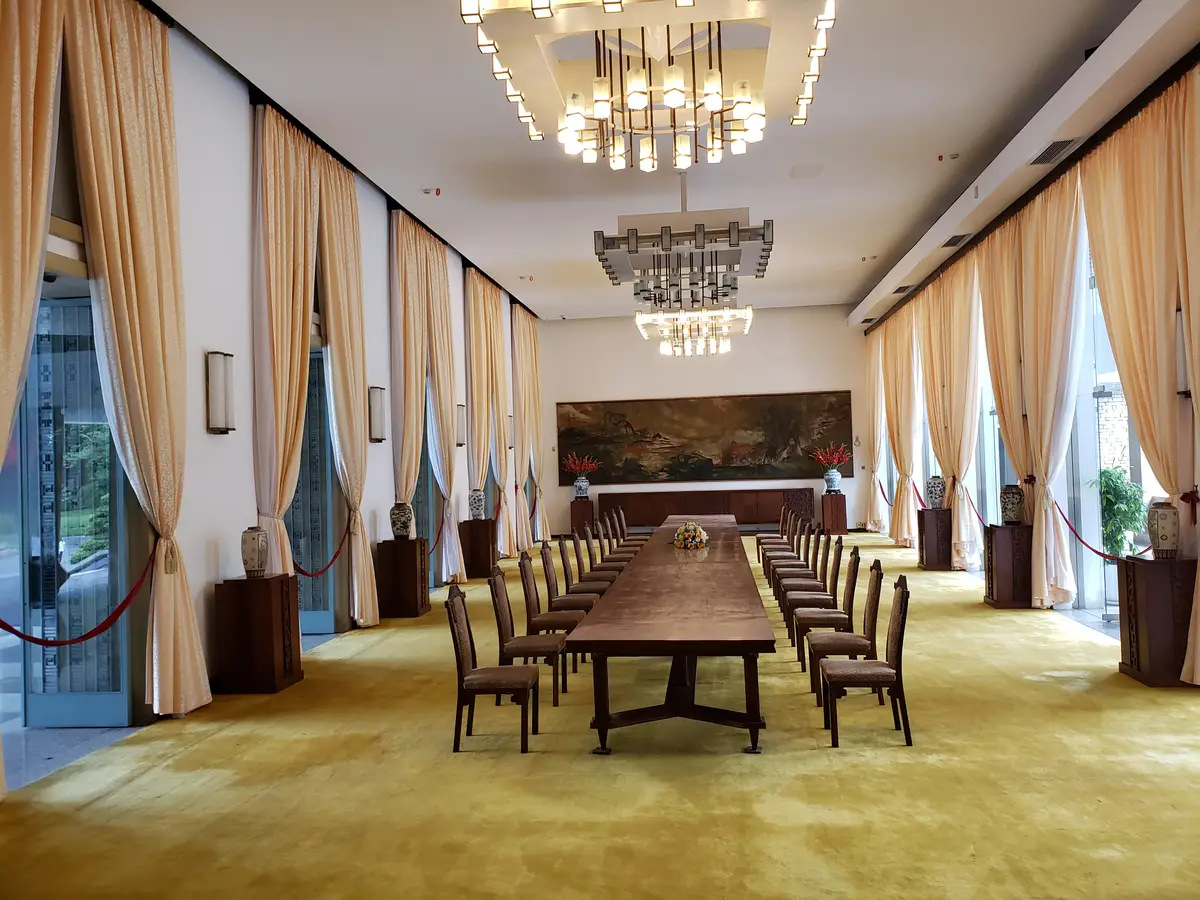 Long wooden meeting table with chairs along both sides in a hall with cream curtains and chandeliers