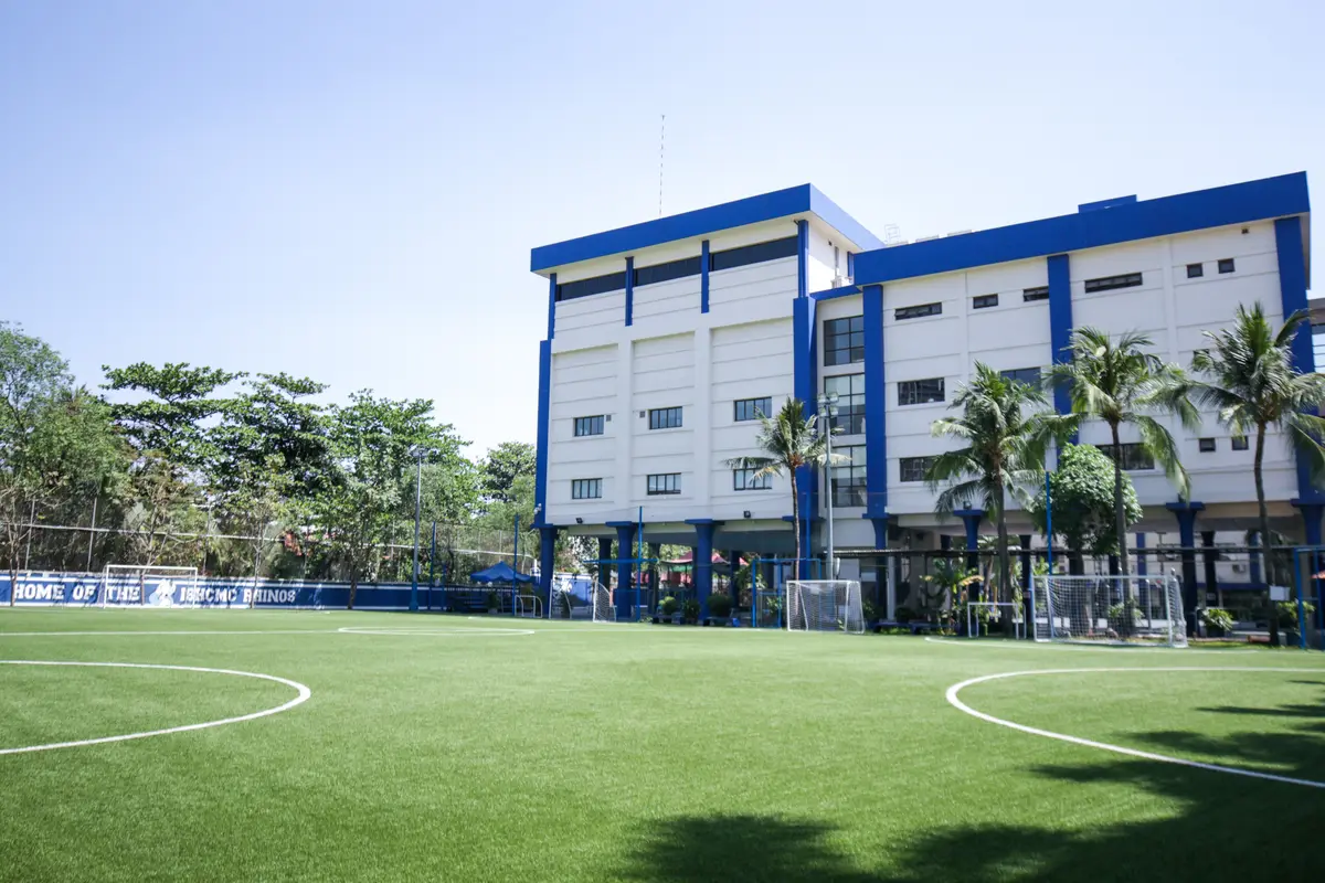 Blue and white school blocks behind a green football pitch and palm trees