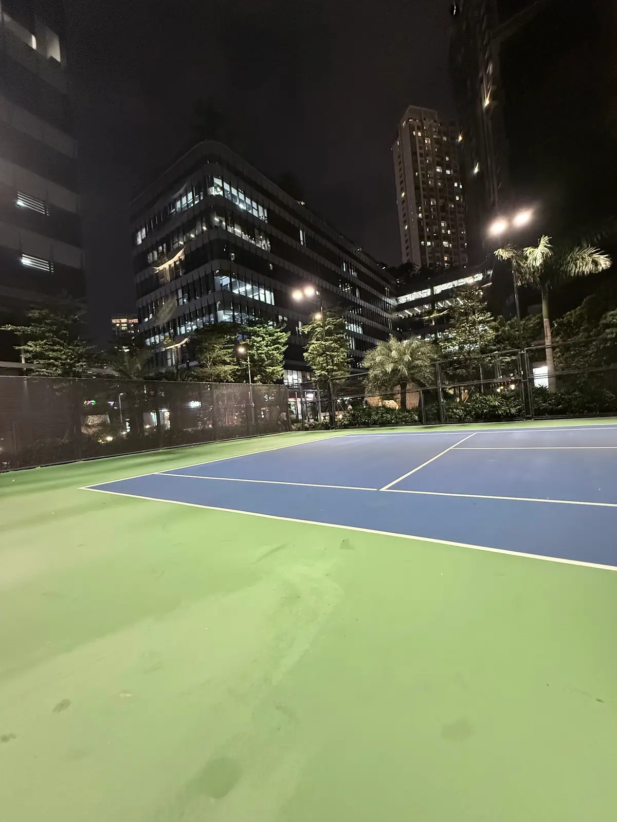 Empty blue hard court with green surround at night in front of tall residential towers