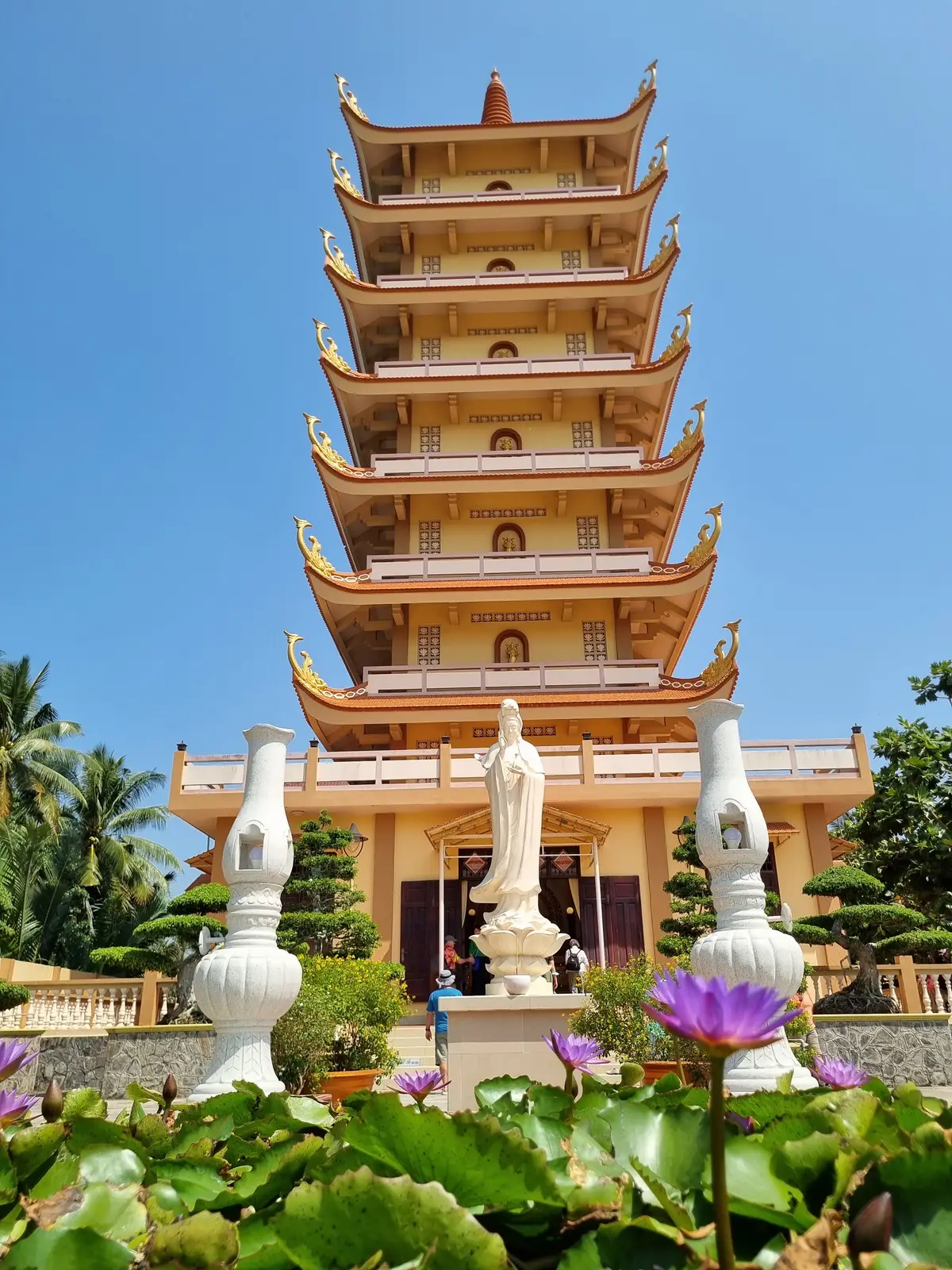 Tiered tower with curved roofs, white statues and palm trees against a blue sky