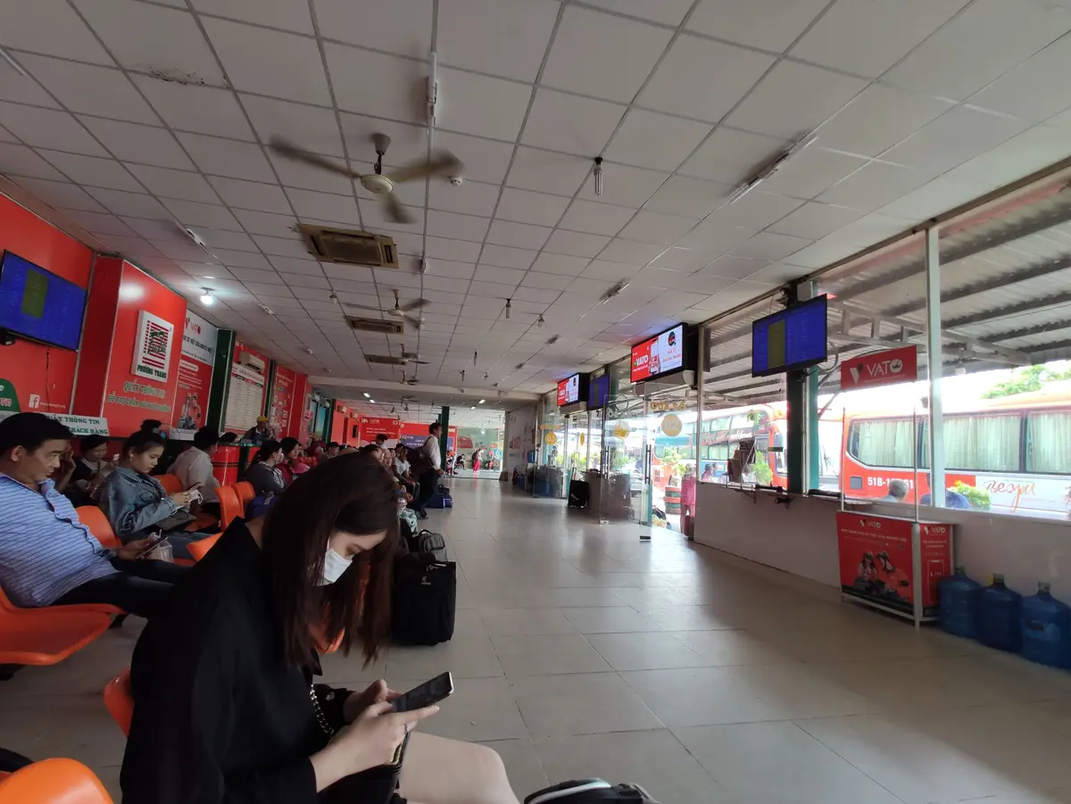 Indoor waiting hall with ticket counters, orange seats and passengers on their phones