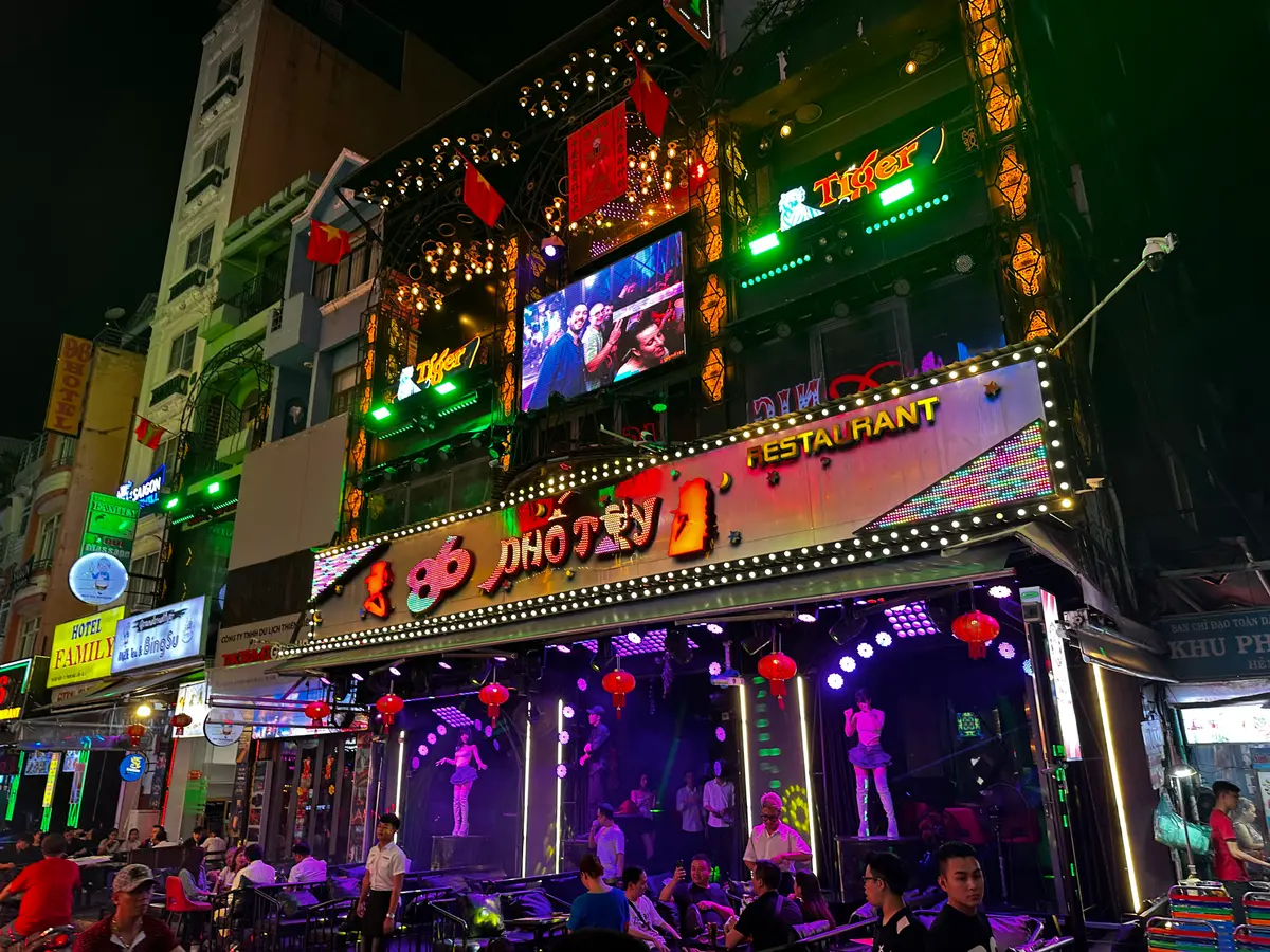 Restaurant and bar frontage covered in neon signs with people seated outside at night