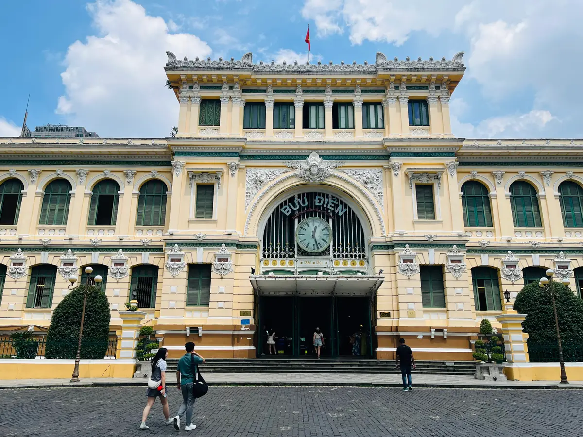 Post office front with a central arched entrance, a large clock and people crossing the paving