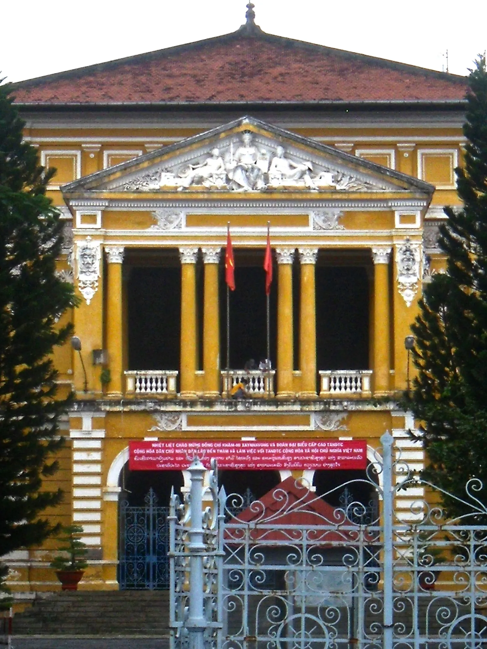 Yellow colonial building with columns, two red flags and a sculpted pediment above an iron gate