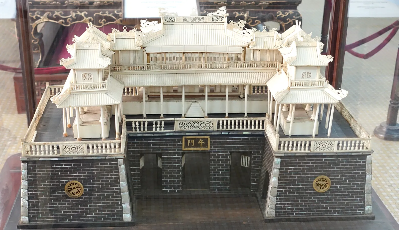 White ivory model of a palace gate with two pavilions on a dark stone base in a glass case