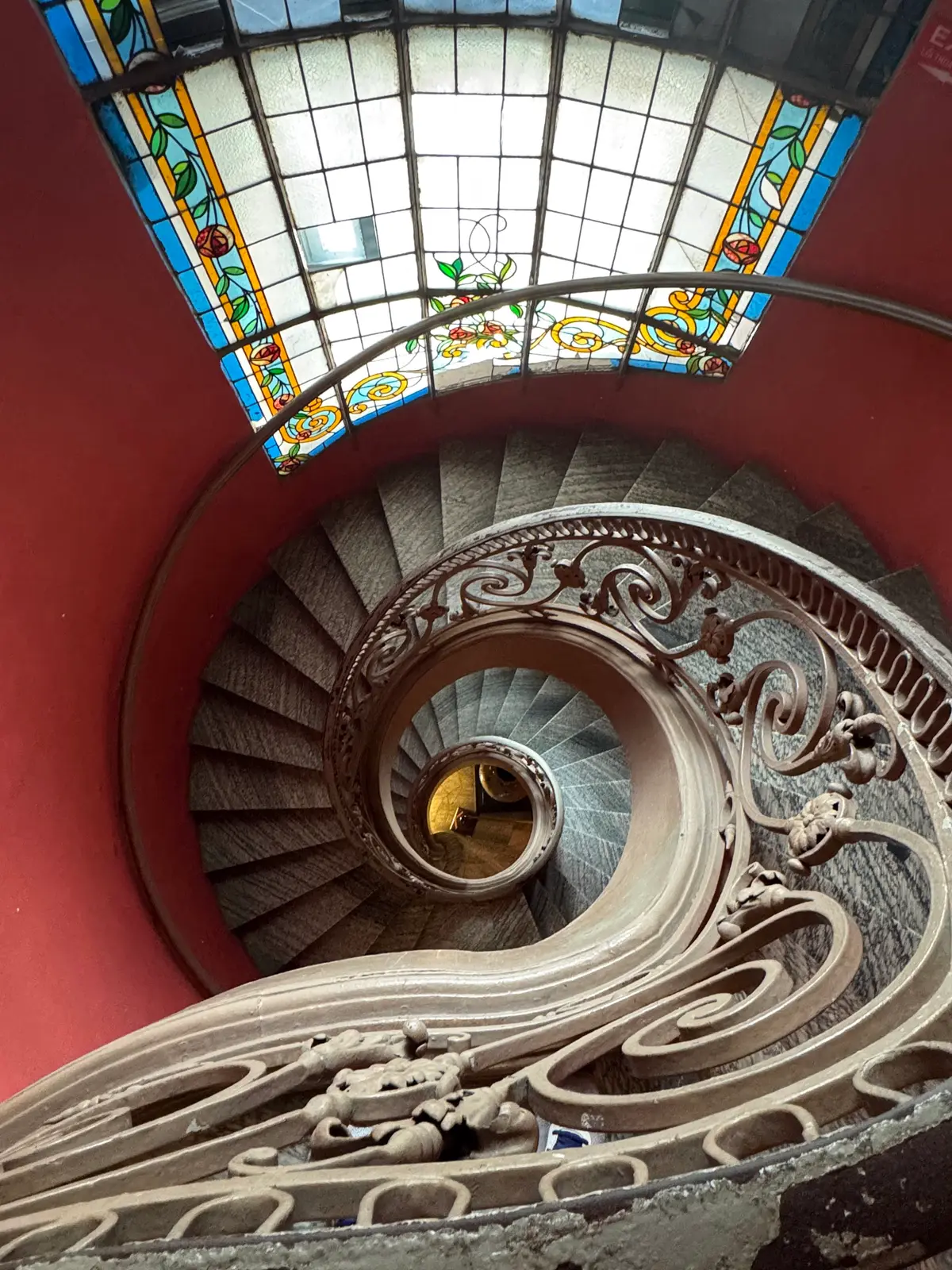 Looking down a curling stone staircase with an ornate railing beneath a stained glass skylight and red walls
