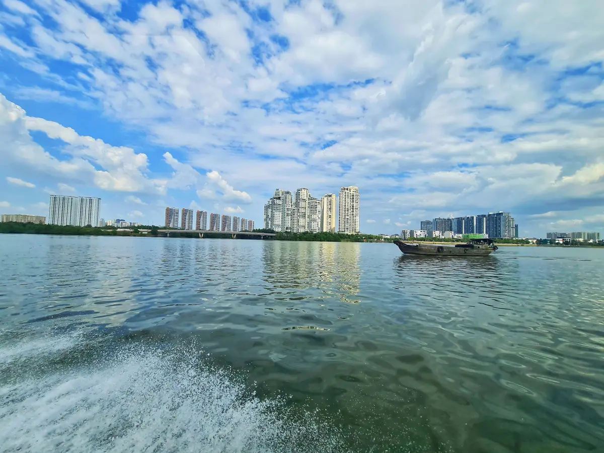 Wake of a boat on a wide green river with a distant bank of apartment blocks under a cloudy sky