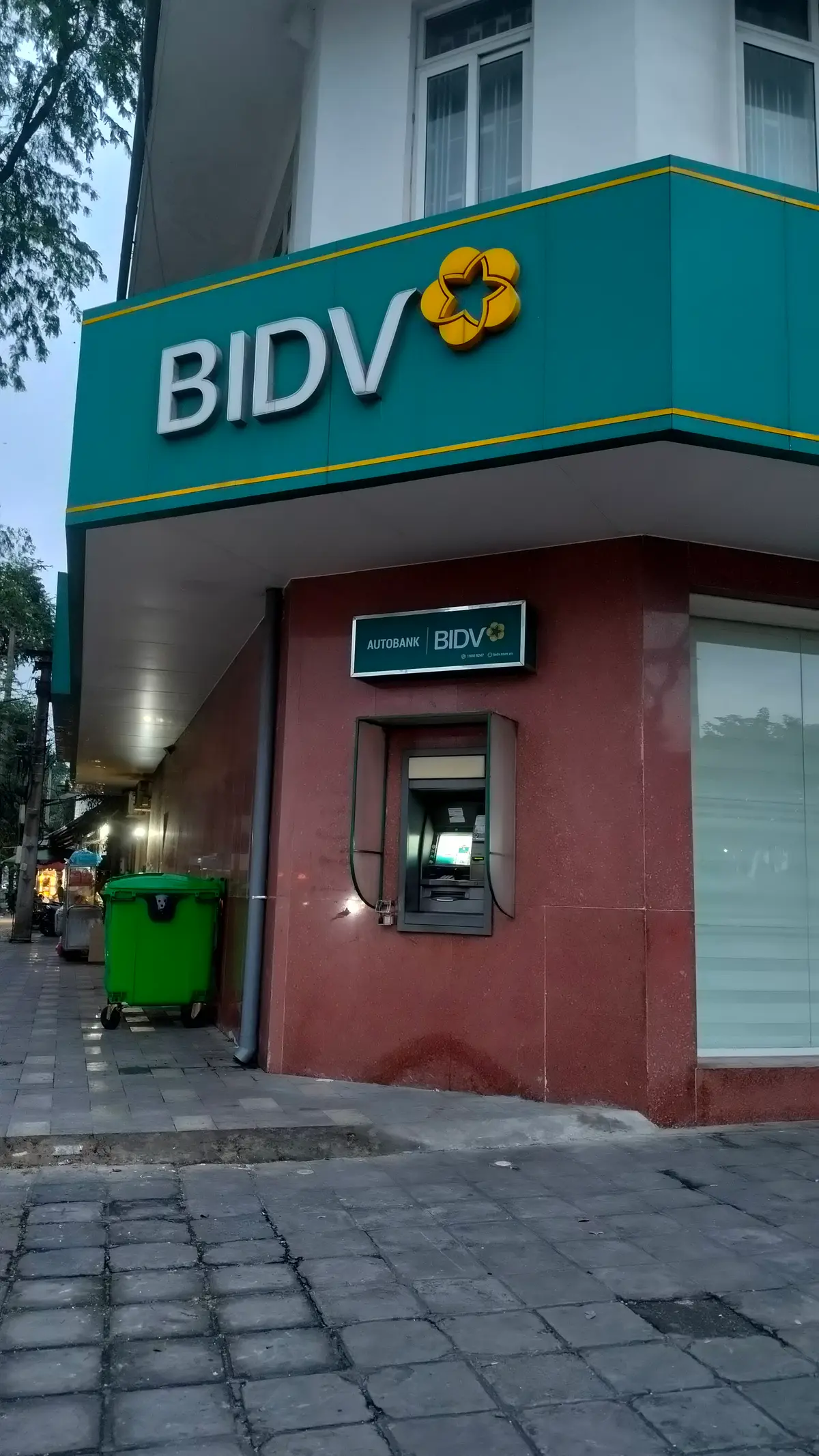 Corner of a bank building with a green BIDV sign above a cash machine set into a dark red wall, a green waste bin on the pavement.
