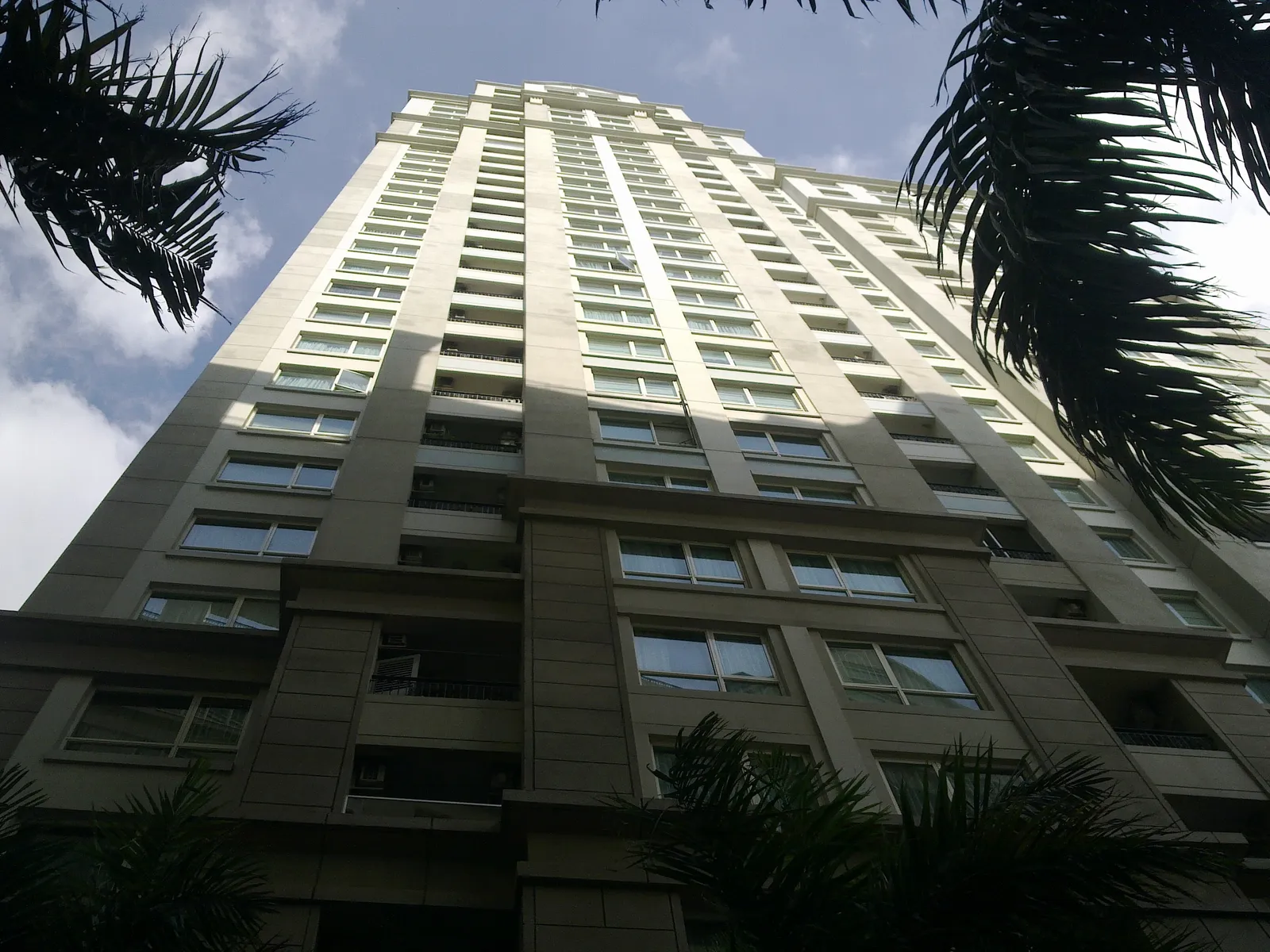 Looking up the side of a tall beige apartment tower between palm leaves