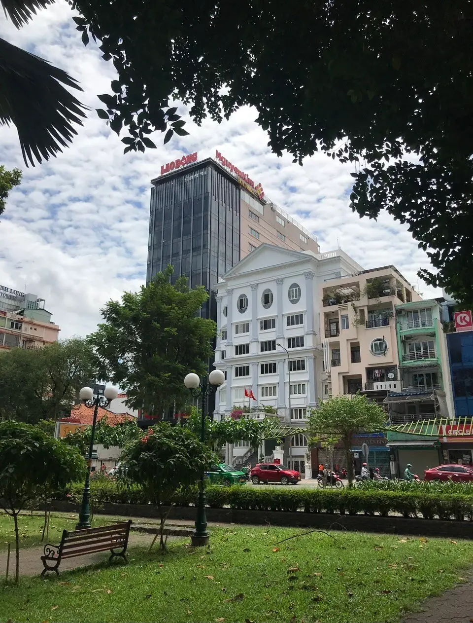 Street scene with a park bench and trees in front of a tall office tower and a white low-rise building.