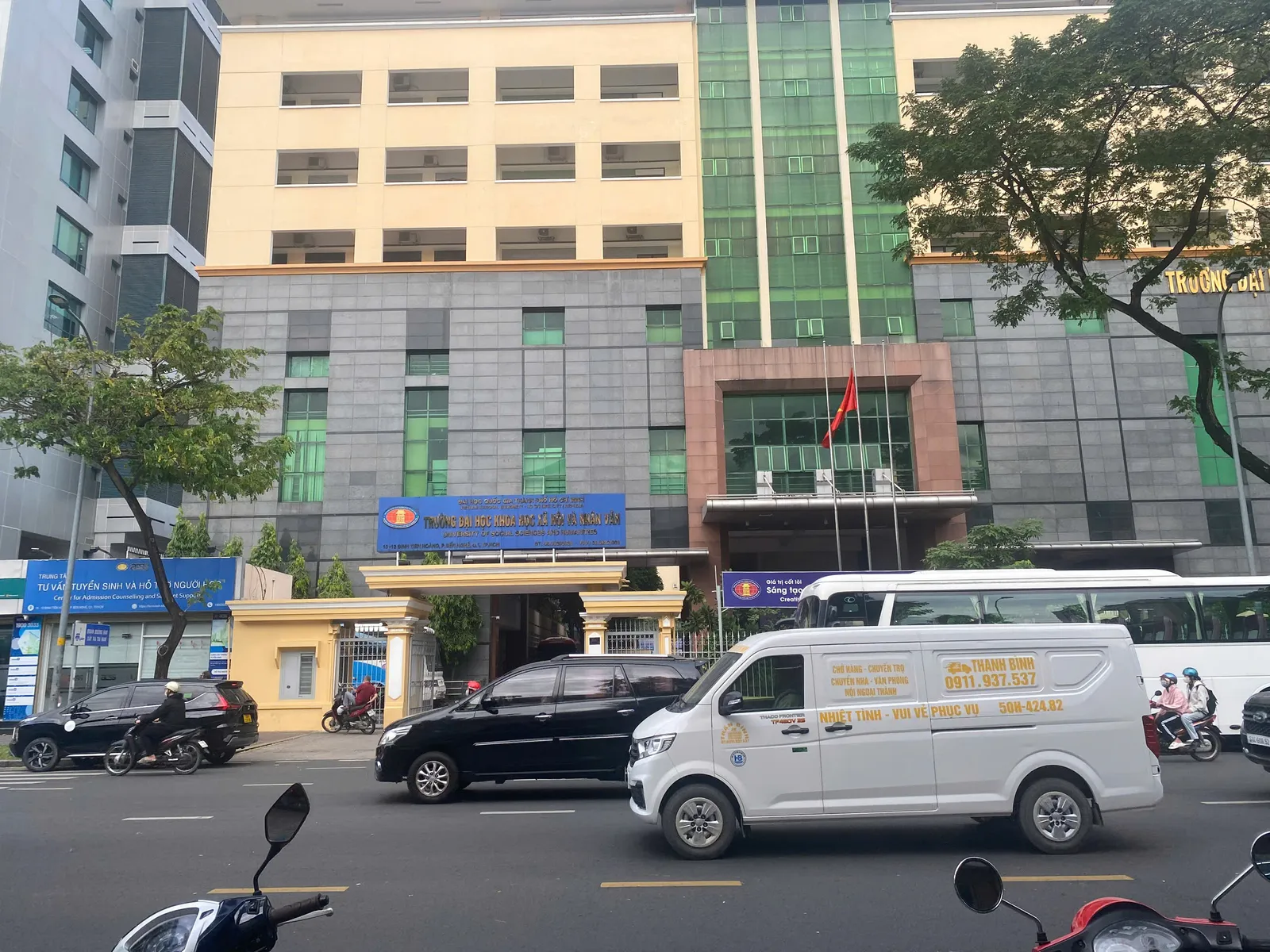 Grey and yellow multi-storey university building with a blue sign above the entrance, a white van and motorbikes on the street in front