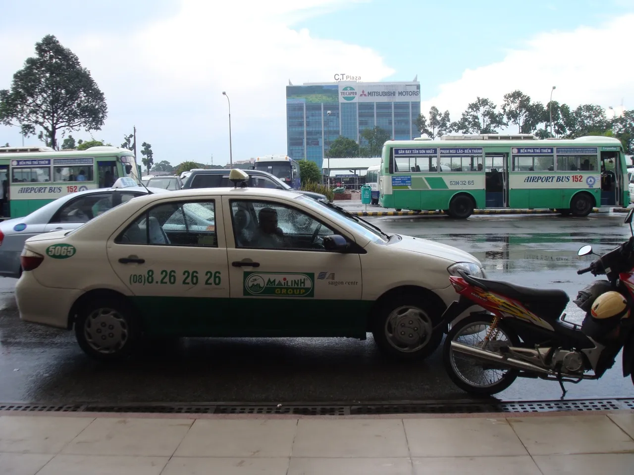 White and green taxi parked at an airport curb with city buses and a motorbike behind it