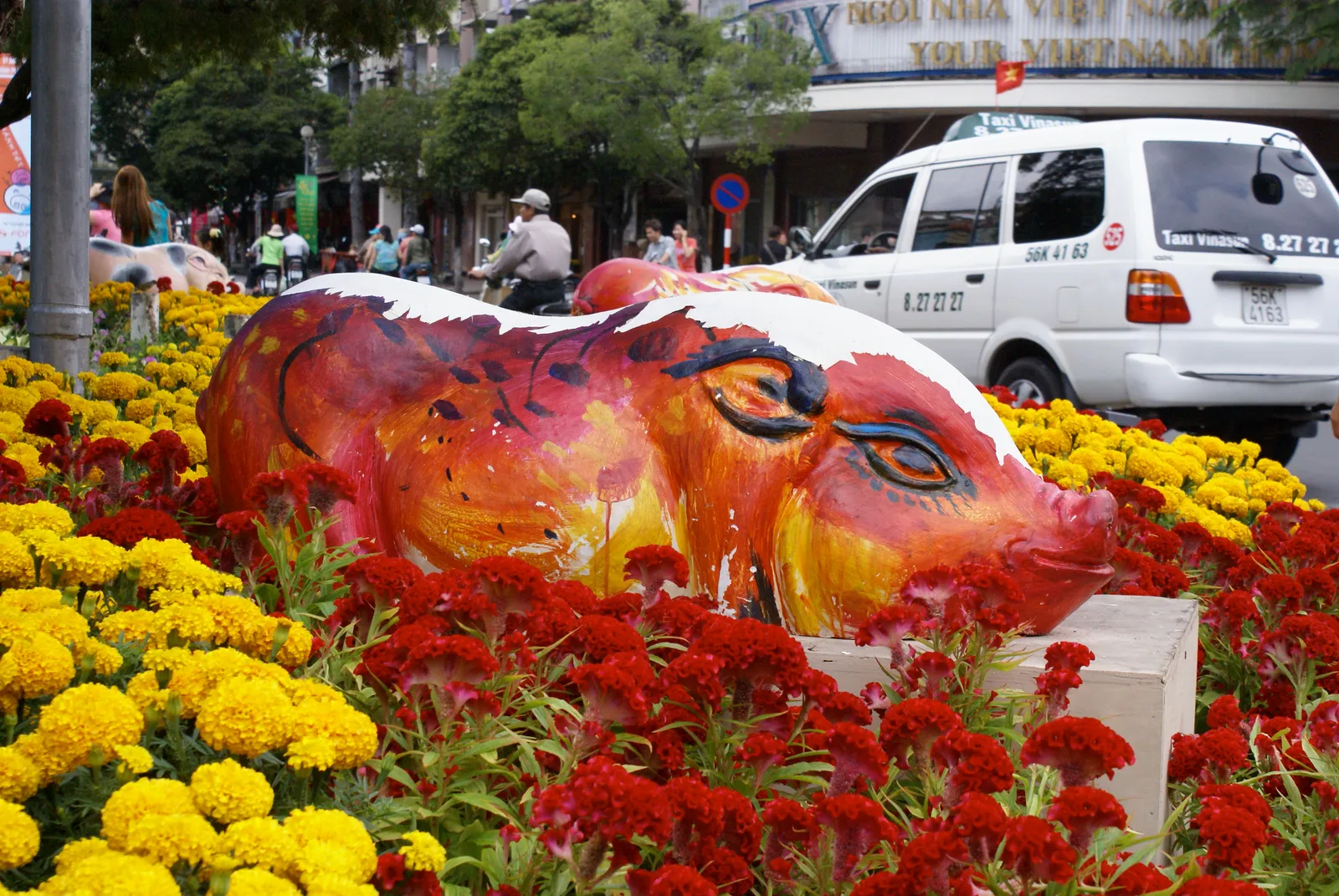 Painted pig sculpture on a white plinth among red and yellow flowers beside a street