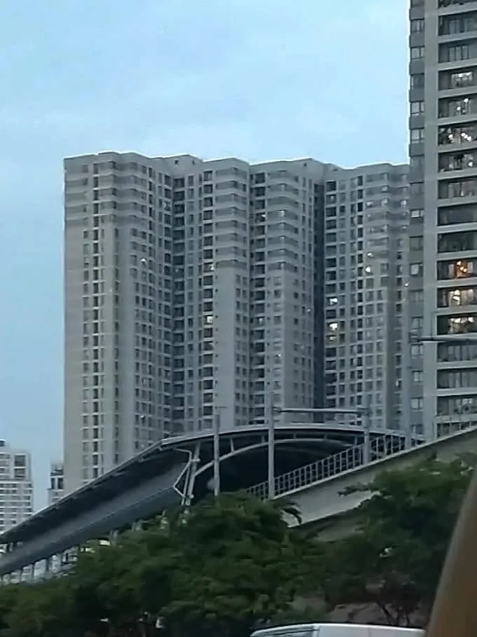 Grey high-rise apartment towers behind trees with a curved elevated metro track below
