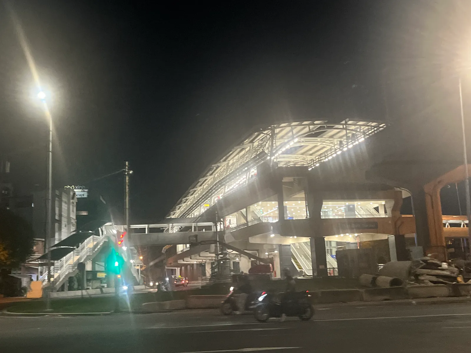 Lit metro station canopy at night beside a road with a traffic light and motorbikes