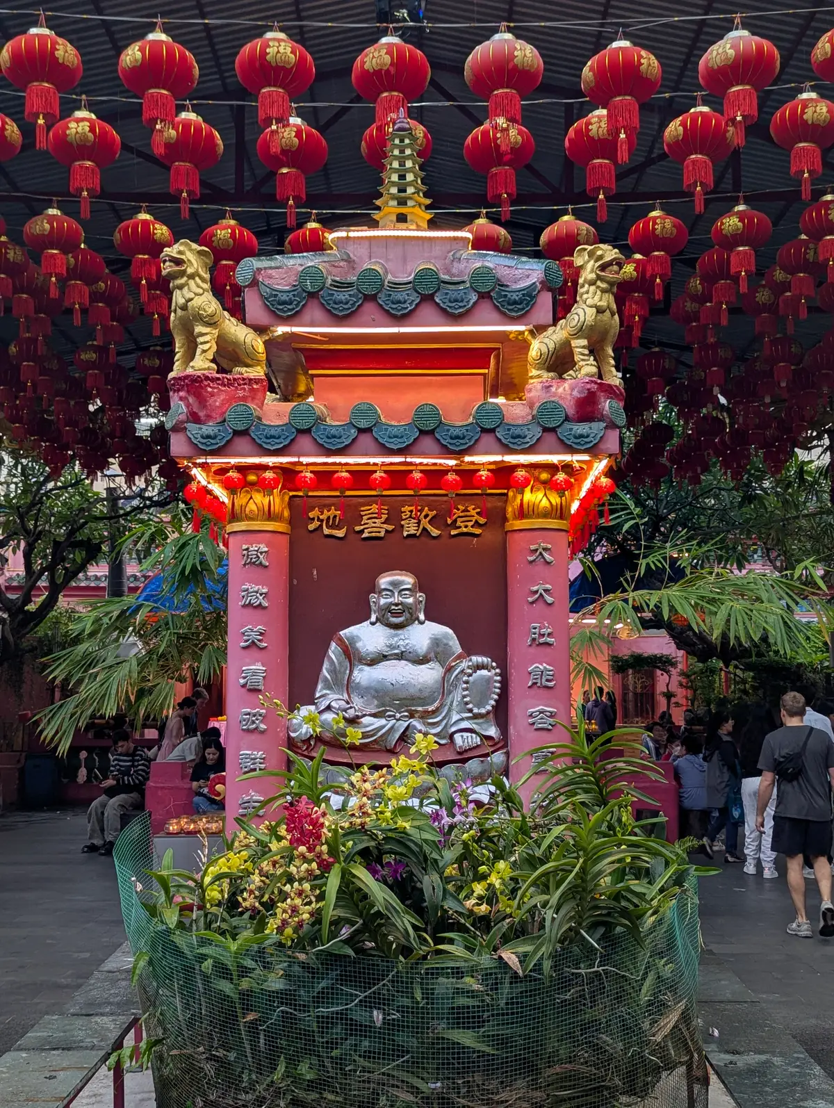 Smiling Buddha statue in a red and pink shrine with lanterns and plants around it