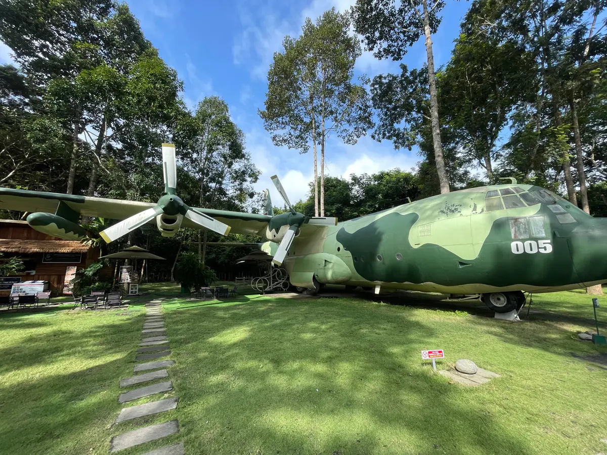 Green military transport aircraft parked on a lawn among tall trees