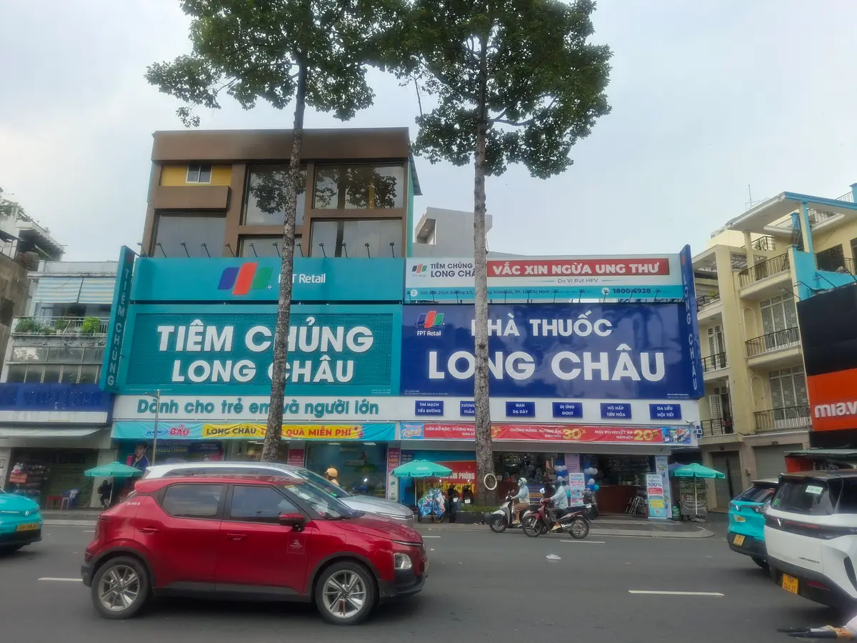 Street view of a row of shopfronts with blue Long Chau pharmacy and vaccination signs.