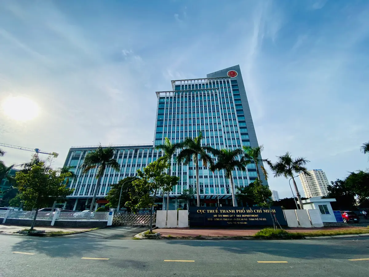 Blue glass office tower with a lower wing, palm trees and a street in front, under a clear sky.