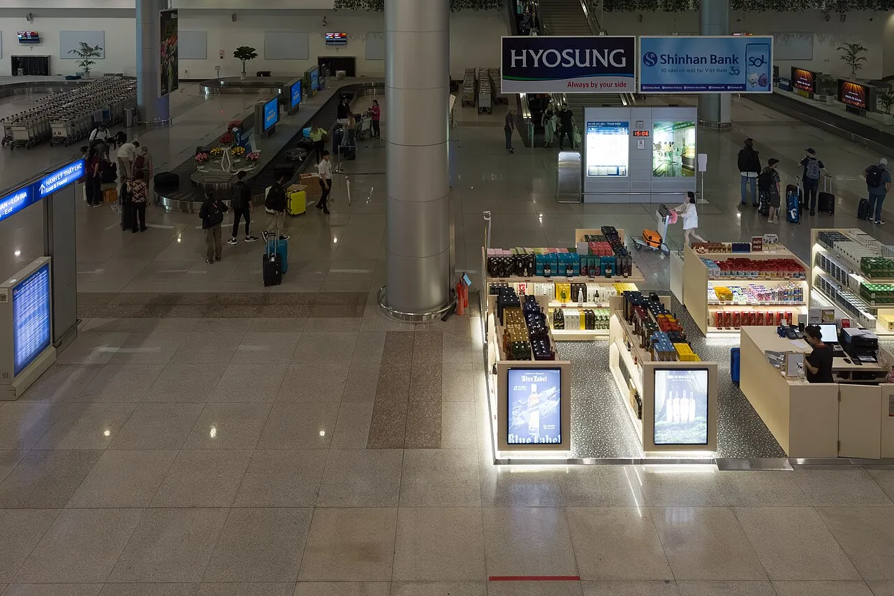 Dim international arrivals hall with a lit duty-free shop, a bank sign overhead and travellers walking across a polished floor.