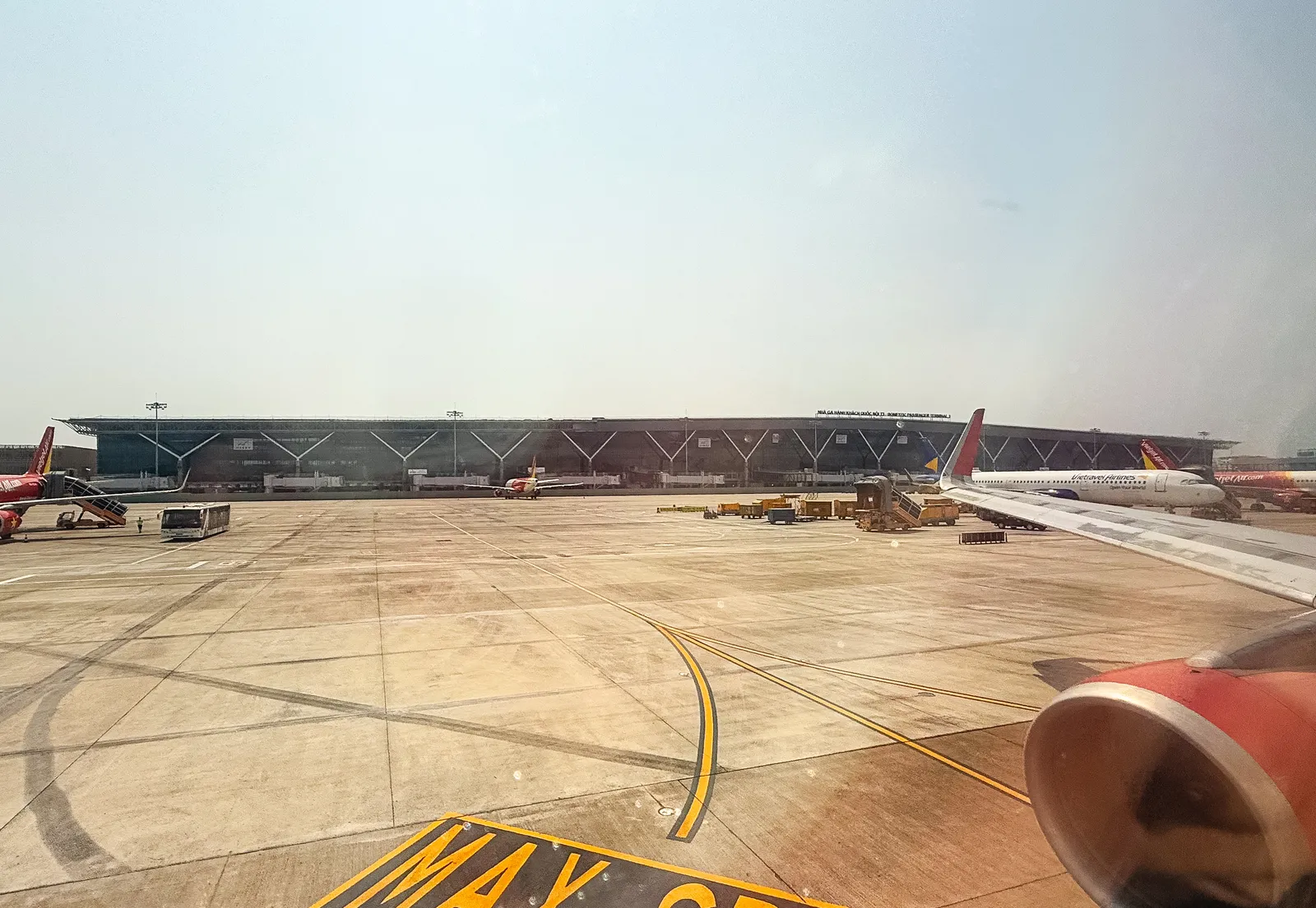 Airport apron with markings and parked aircraft in front of a long terminal building, seen through an aircraft window