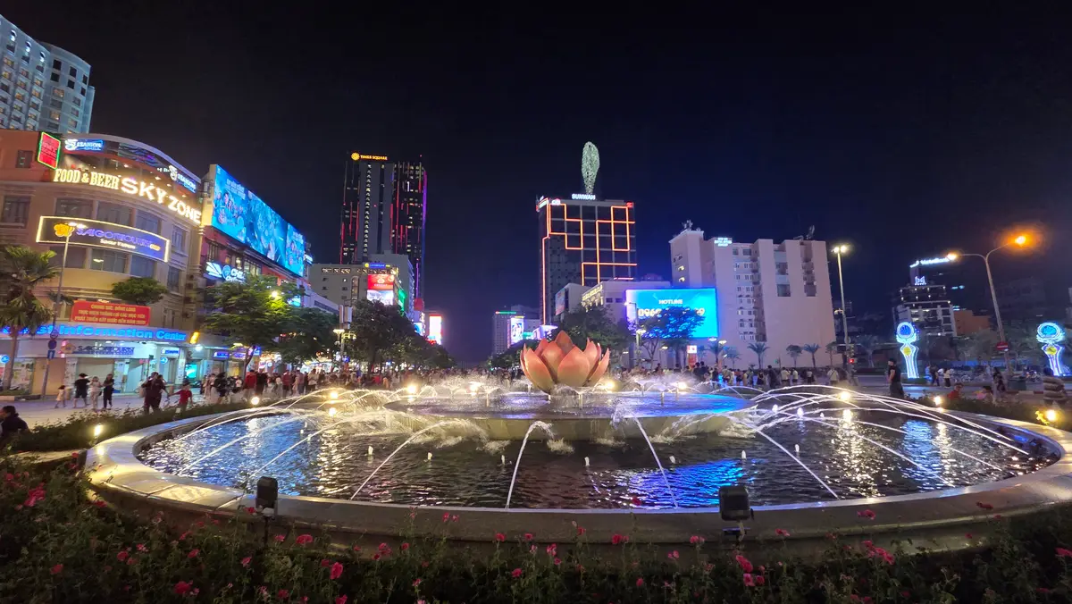 Lit fountain with a lotus-shaped centre on a street at night with bright signs behind